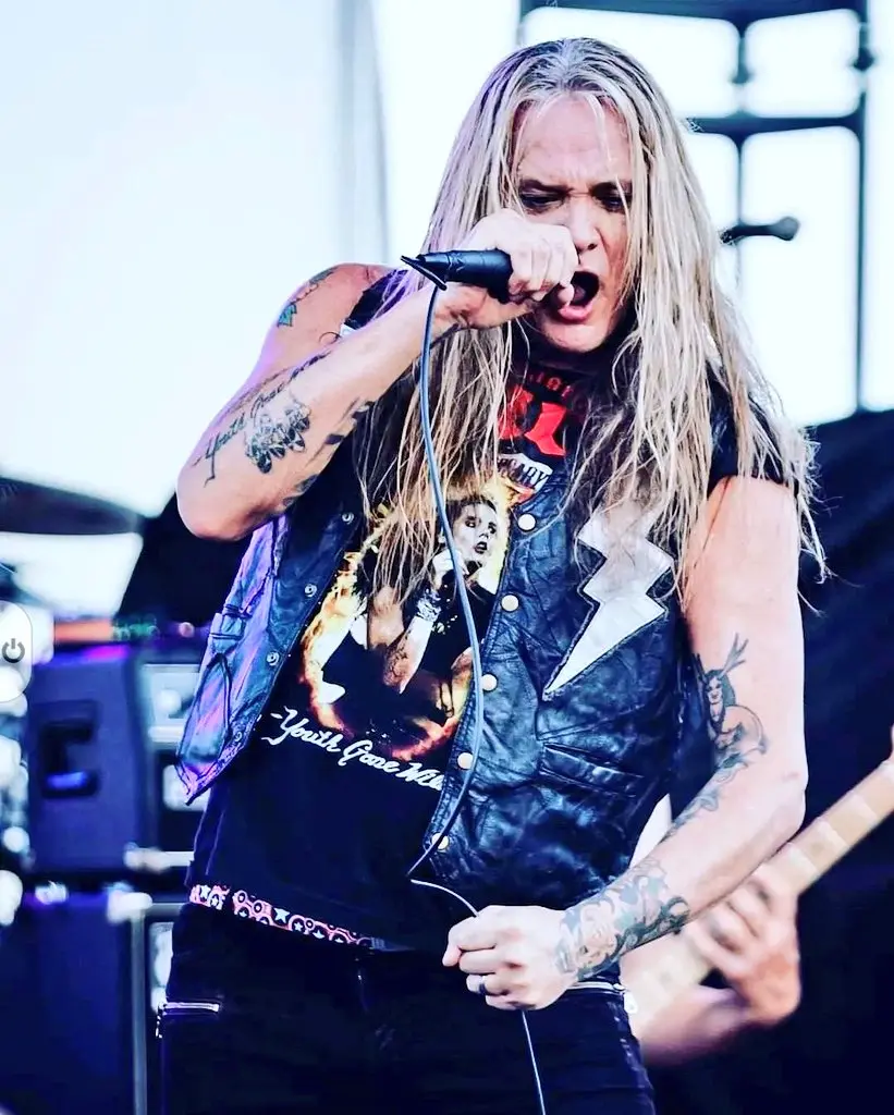 The image shows Sebastian Bach performing on stage with long hair, a leather vest, and a rock t-shirt.